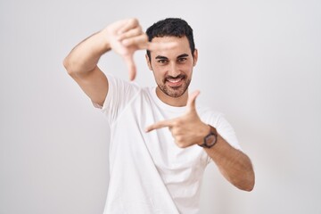 Handsome hispanic man standing over white background smiling making frame with hands and fingers with happy face. creativity and photography concept.
