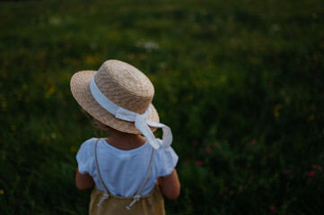 Rear view of adorable little girl with straw hat standing in the middle of summer meadow.