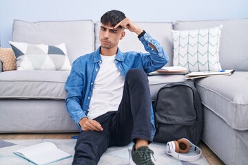Young hispanic man sitting on the floor studying for university pointing unhappy to pimple on...