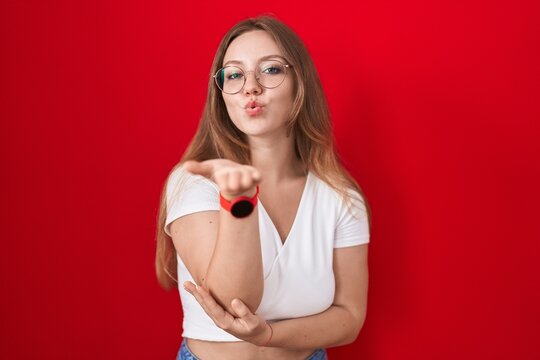 Young caucasian woman standing over red background looking at the camera blowing a kiss with hand on air being lovely and sexy. love expression.