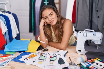 Young beautiful hispanic woman tailor smiling confident sitting on table at tailor shop