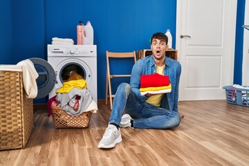 Young hispanic man holding clean laundry in shock face, looking skeptical and sarcastic, surprised...