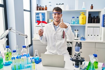Young hispanic man working at scientist laboratory holding diploma smiling happy and positive, thumb up doing excellent and approval sign