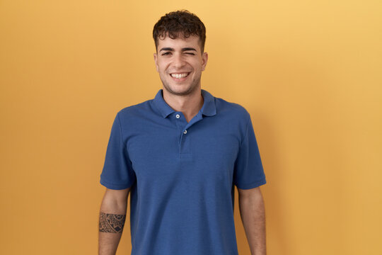 Young hispanic man standing over yellow background winking looking at the camera with sexy expression, cheerful and happy face.