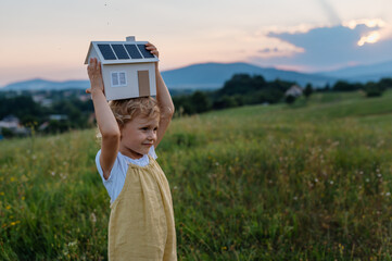 Little girl with model house with installed of solar panels, standing in the middle of meadow.