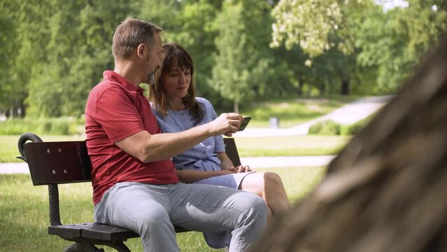 A middle-aged Caucasian couple laughs and looks at a smartphone as they sit on a bench in a park on a sunny day
