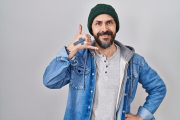 Young hispanic man with tattoos wearing wool cap smiling doing phone gesture with hand and fingers like talking on the telephone. communicating concepts.