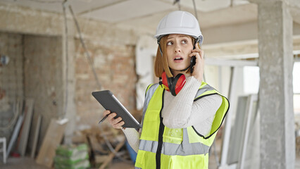 Young blonde woman architect talking on smartphone looking touchpad at construction site