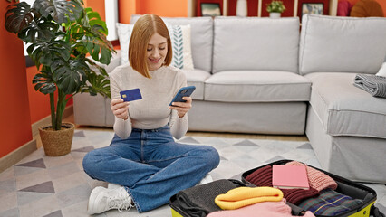 Young blonde woman shopping flight with smartphone and credit card sitting on floor at home