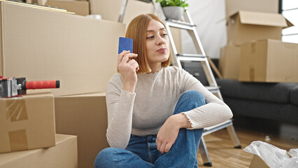 Young blonde woman sitting on floor thinking holding credit card at new home
