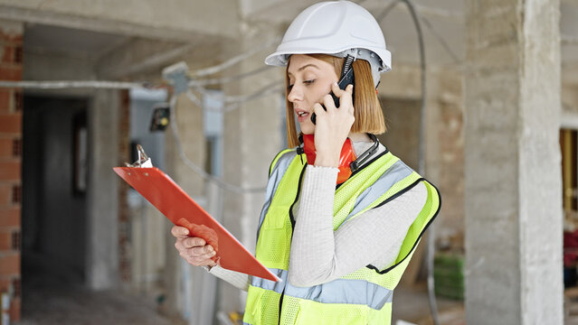 Young blonde woman architect talking on smartphone reading document at construction site