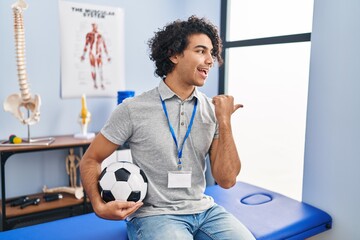 Hispanic man with curly hair working as football physiotherapist smiling with happy face looking and pointing to the side with thumb up.