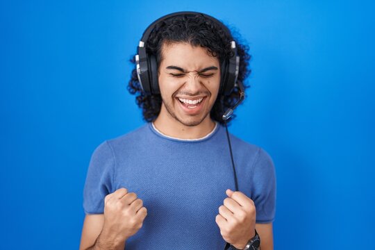 Hispanic man with curly hair listening to music using headphones very happy and excited doing winner gesture with arms raised, smiling and screaming for success. celebration concept.