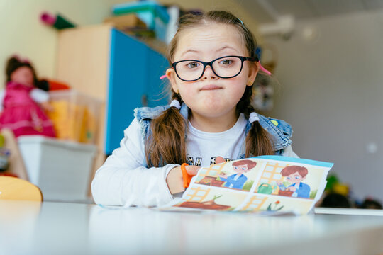 Cute little junior schoolchild with special needs having funny emotion on her face sits at desk at classroom works independently.