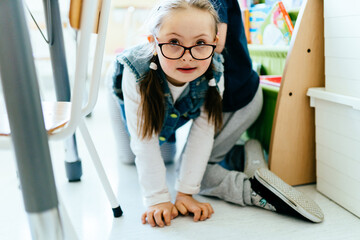 Smiling junior cauasian school girl with down syndrome crawling on the floor in the classroom playing with children.