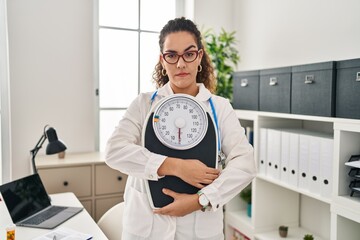 Young hispanic woman working at dietitian clinic skeptic and nervous, frowning upset because of...