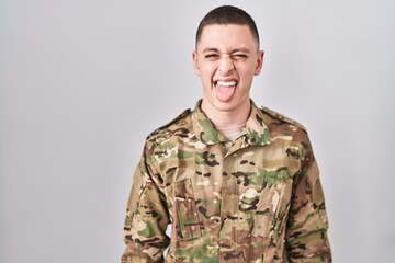 Young man wearing camouflage army uniform sticking tongue out happy with funny expression. emotion concept.