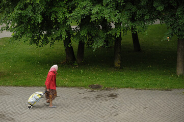 an old woman in a headscarf in a red sweater drags a cart down the street