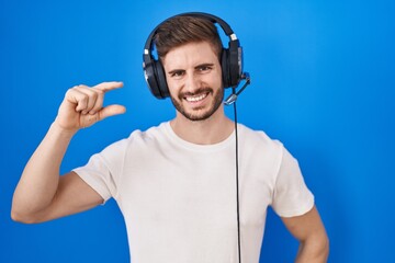 Hispanic man with beard listening to music wearing headphones smiling and confident gesturing with hand doing small size sign with fingers looking and the camera. measure concept.