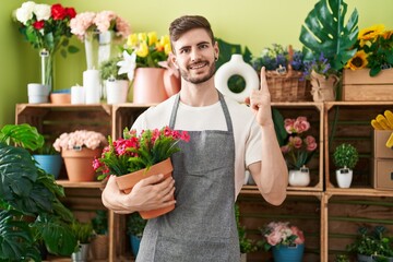 Hispanic man with beard working at florist shop holding plant smiling with an idea or question pointing finger with happy face, number one