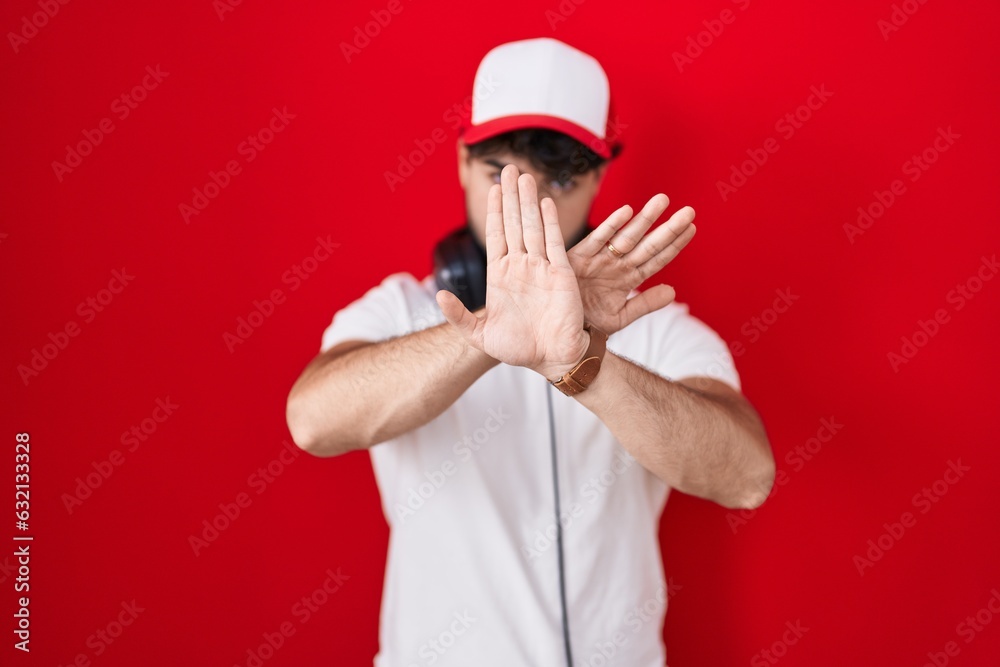 Poster Hispanic man with beard wearing gamer hat and headphones rejection expression crossing arms and palms doing negative sign, angry face