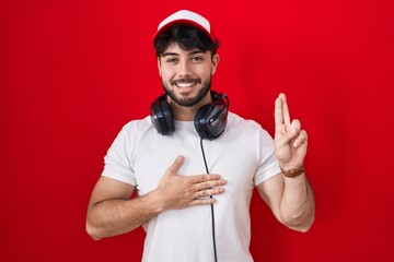 Hispanic man with beard wearing gamer hat and headphones smiling swearing with hand on chest and...