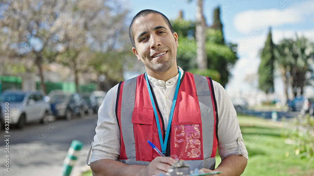 Wall mural Young hispanic man volunteer smiling holding clipboard at park