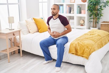 Young latin man suffering for stomach ache sitting on bed at bedroom