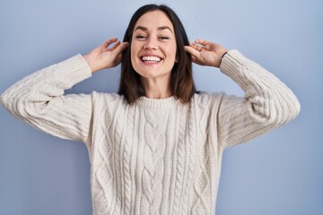 Young brunette woman standing over blue background smiling pulling ears with fingers, funny...