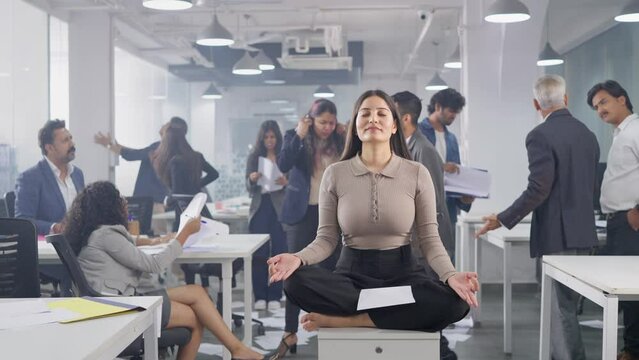 A young Asian Indian corporate female executive sitting quietly eyes closed in a meditative pose middle of a busy chaotic and messy office where people or coworkers throw papers, fight, and shout. 