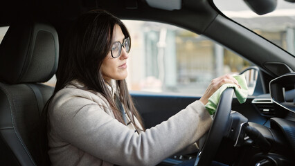 Young beautiful hispanic woman cleaning steering wheel sitting on car at street