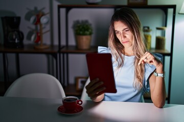 Young hispanic woman using touchpad sitting on the table at night pointing down looking sad and upset, indicating direction with fingers, unhappy and depressed.