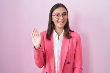 Young hispanic woman wearing business clothes and glasses waiving saying hello happy and smiling,...