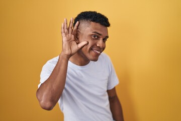 Young hispanic man standing over yellow background smiling with hand over ear listening an hearing to rumor or gossip. deafness concept.