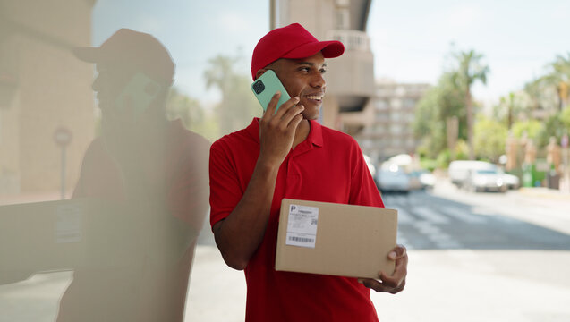 Young latin man delivery worker holding package talking on smartphone at street