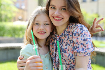 A happy mother and daughter hugging and smiling in a park. They wear green and patterned shirts and have fun with a straw and a peace sign.