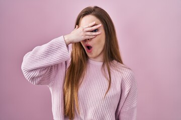 Young caucasian woman standing over pink background peeking in shock covering face and eyes with hand, looking through fingers with embarrassed expression.