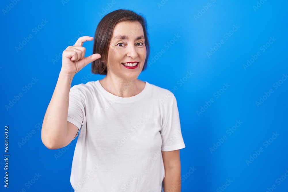 Poster Middle age hispanic woman standing over blue background smiling and confident gesturing with hand doing small size sign with fingers looking and the camera. measure concept.