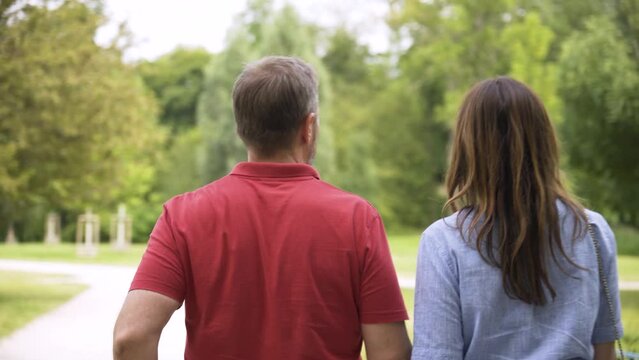 A middle-aged Caucasian couple walks through a park on a sunny day - view from behind