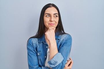 Hispanic woman standing over blue background looking confident at the camera smiling with crossed...