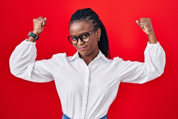 African woman with braids standing over red background showing arms muscles smiling proud. fitness...