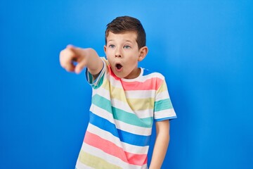 Young caucasian kid standing over blue background pointing with finger surprised ahead, open mouth amazed expression, something on the front
