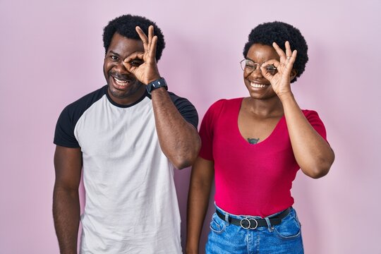 Young african american couple standing over pink background doing ok gesture with hand smiling, eye looking through fingers with happy face.