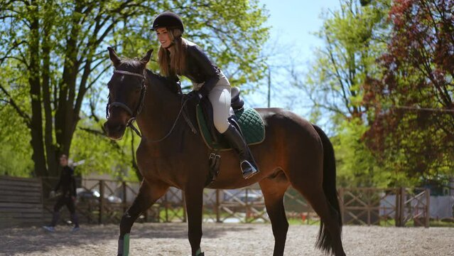 Side view tracking shot of happy woman riding horse in sunshine leaving. Live camera follows satisfied female Caucasian gorgeous equestrian on back of purebred stallion outdoors