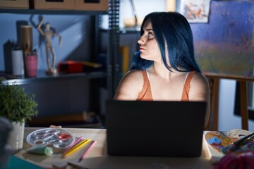 Fototapeta na wymiar Young modern girl with blue hair sitting at art studio with laptop at night looking to side, relax profile pose with natural face with confident smile.
