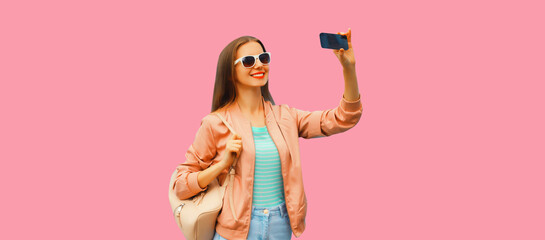 Portrait of happy smiling teenage girl taking selfie with smartphone wearing backpack on pink background