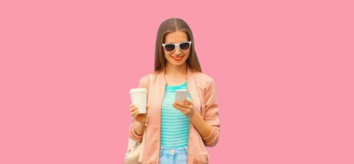 Portrait of young woman with smartphone and cup of coffee wearing sunglasses on pink background