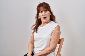 Middle age hispanic woman getting vaccine showing arm with band aid in shock face, looking...