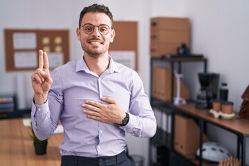 Young hispanic man at the office smiling swearing with hand on chest and fingers up, making a loyalty promise oath