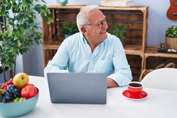 Senior grey-haired man using laptop and drinking coffee sitting on table at home